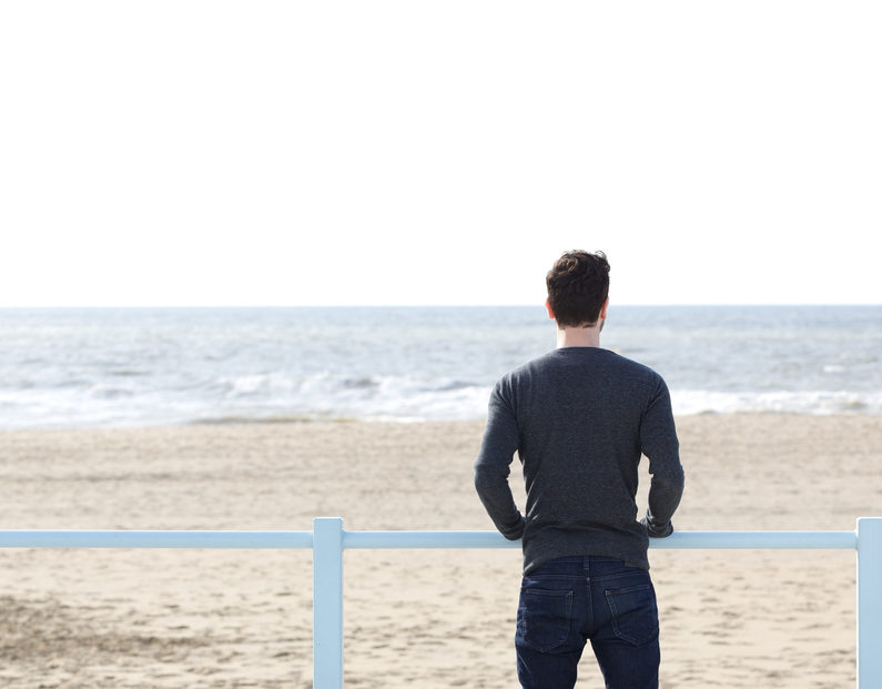 Portrait of a young man standing alone outdoors looking at sea - from behind